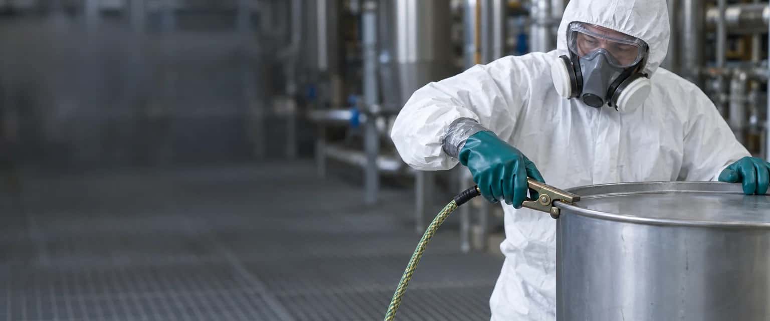 Aftapplek in een chemische installatie met roestvrijstalen leidingen. Rechts klemt iemand in een witte wegwerpoverall met capuchon, ruimzichtbril, halfgelaatsmasker en groene chemicaliënhandschoenen een messing aardingsklem met gevlochten kabel op de rand van een blanco stalen vat.