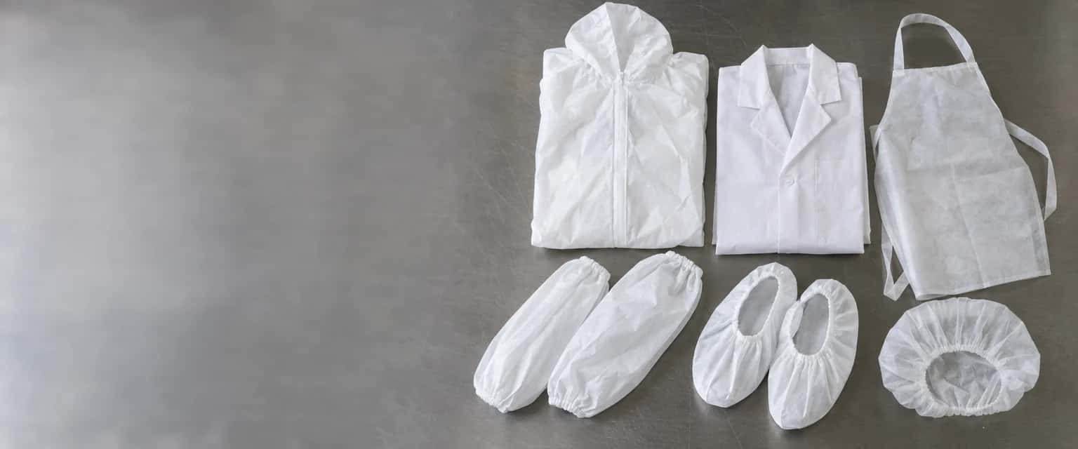 Roestvrijstalen werkblad. Rechts ligt de wegwerpkleding naast elkaar uitgelegd: een opgevouwen witte overall met capuchon, een labjas, een schort, een paar overmouwen, een paar overschoenen en een muts, alle van wit vlies en zonder opdruk.